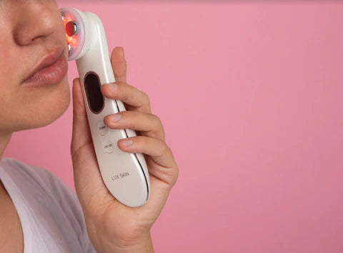 A person using a facial device with a red light on their cheek against a pink background.